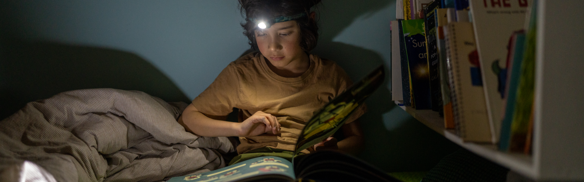 Das Bild zeigt einen Jungen, der im Bett mit einer  Stirnlampe ein Buch liest